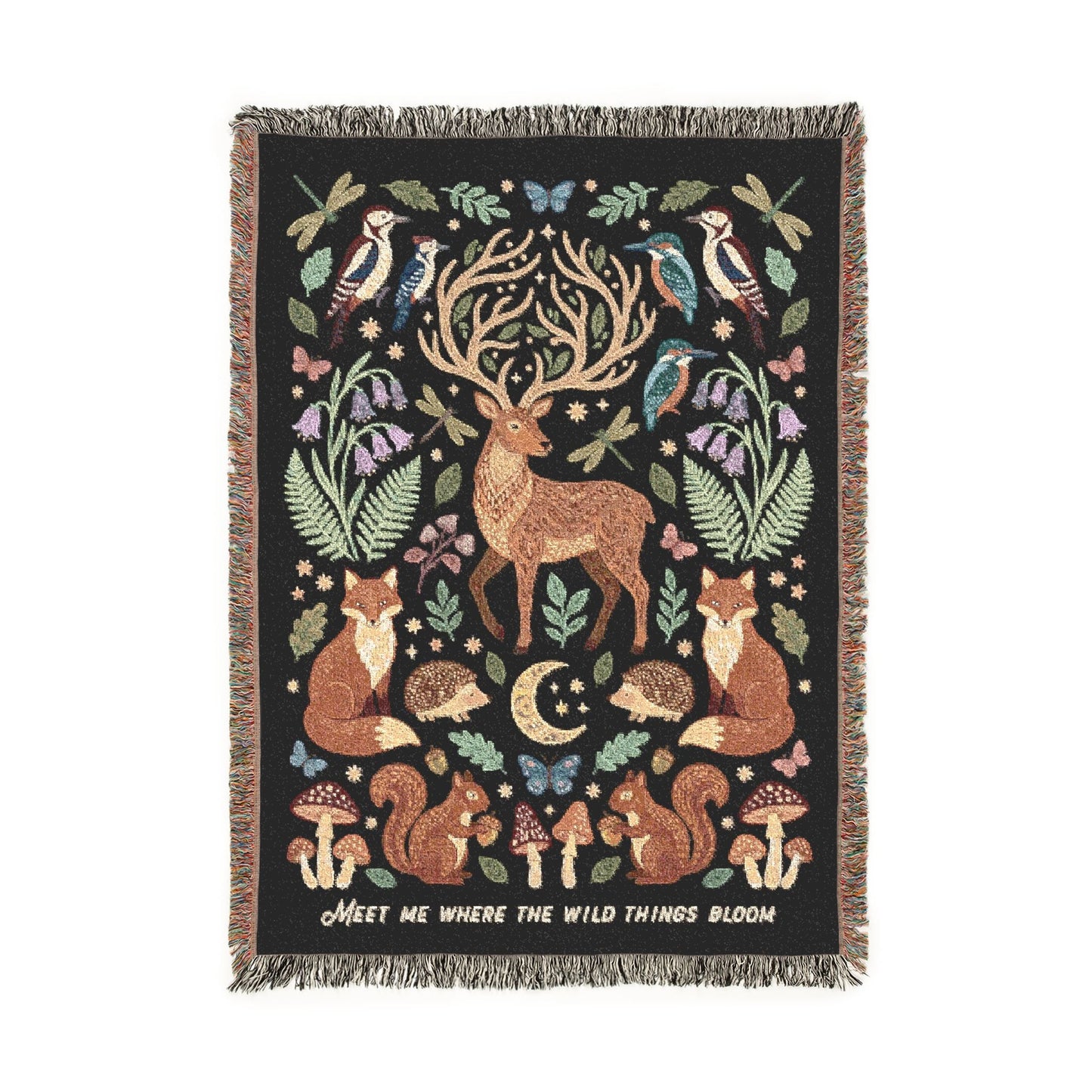 A rectangular woven throw blanket with fringe on all sides, featuring a woodland scene with a stag, foxes, deer, birds, mushrooms, ferns, and a crescent moon on a black background. White text along the right edge reads, MeET ME WHERE THE WILD THINGS BLOOM.