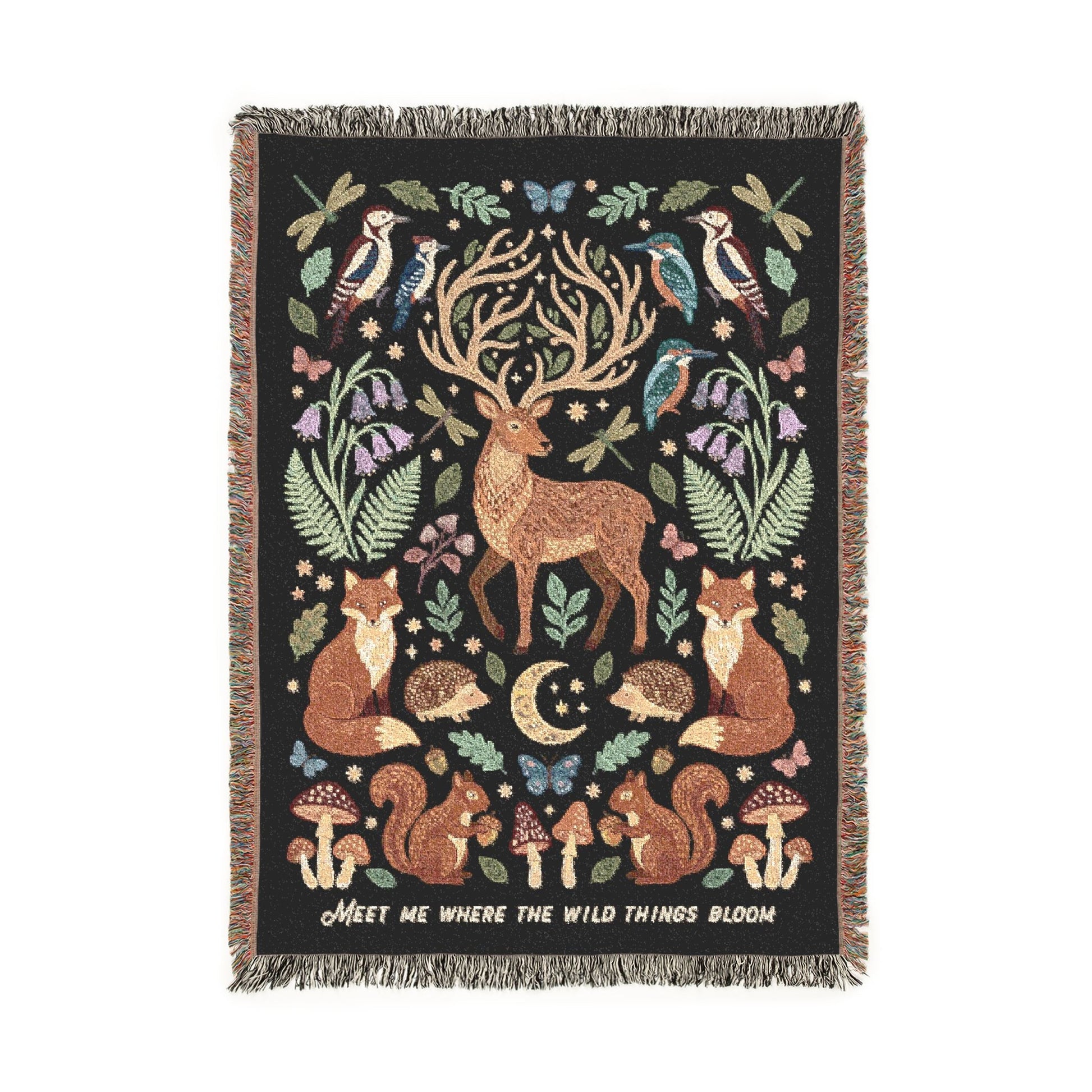 A rectangular woven throw blanket with fringe on all sides, featuring a woodland scene with a stag, foxes, deer, birds, mushrooms, ferns, and a crescent moon on a black background. White text along the right edge reads, MeET ME WHERE THE WILD THINGS BLOOM.